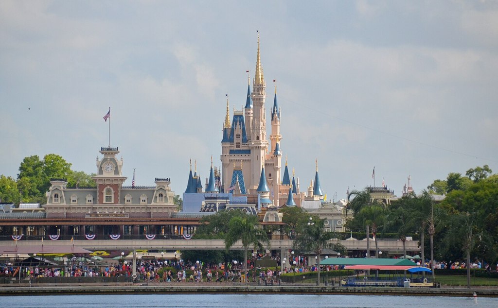 A view of the "Magic Kingdom" in Walt Disney World in Orlando Florida. A fairytale castle's spires rise above a 19th-century mansard roof style town hall, in the foreground there is a fence with patriotic buntin and dozens of people, on the border of a body of water.
Clément Bardot, English:  Magic Kingdom, Disney World, Orlando, Florida, USA, May 27, 2015, May 27, 2015, Own work, https://commons.wikimedia.org/wiki/File:Magic_Kingdom,_Disney_World.jpg#/media/File:Magic_Kingdom,_Disney_World.jpg.